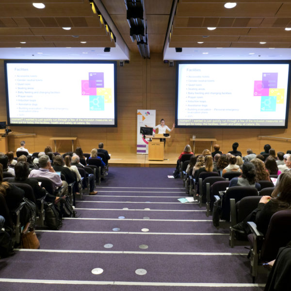 A photo of the 2019 EDIS symposium in the Francis Crick Institute auditorium, showing someone speaking in front of a crowed with the EDIS logo on the display screens behind them
