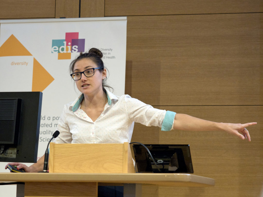 Lilian Hunt, a white non-binary person with long dark hair, speaking at the EDIS 2019 symposium on a stage.