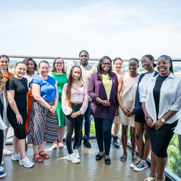A photo of The Equal Group team standing on a balcony in the sun. 15 people smile at the camera, they are a mixture of ethnicities, genders, and ages.